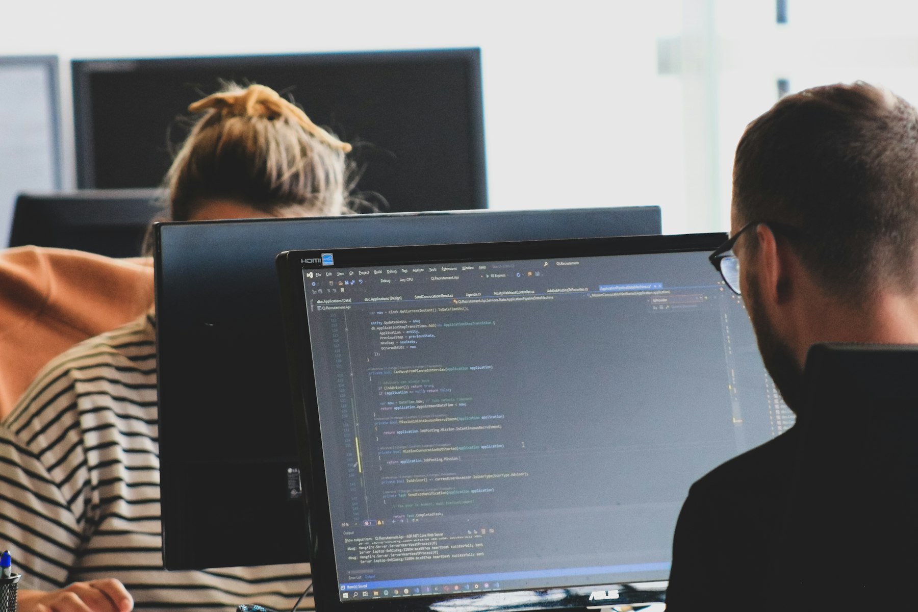 Zwei Personen arbeiten an Monitoren mit Code in einem hellen Büro